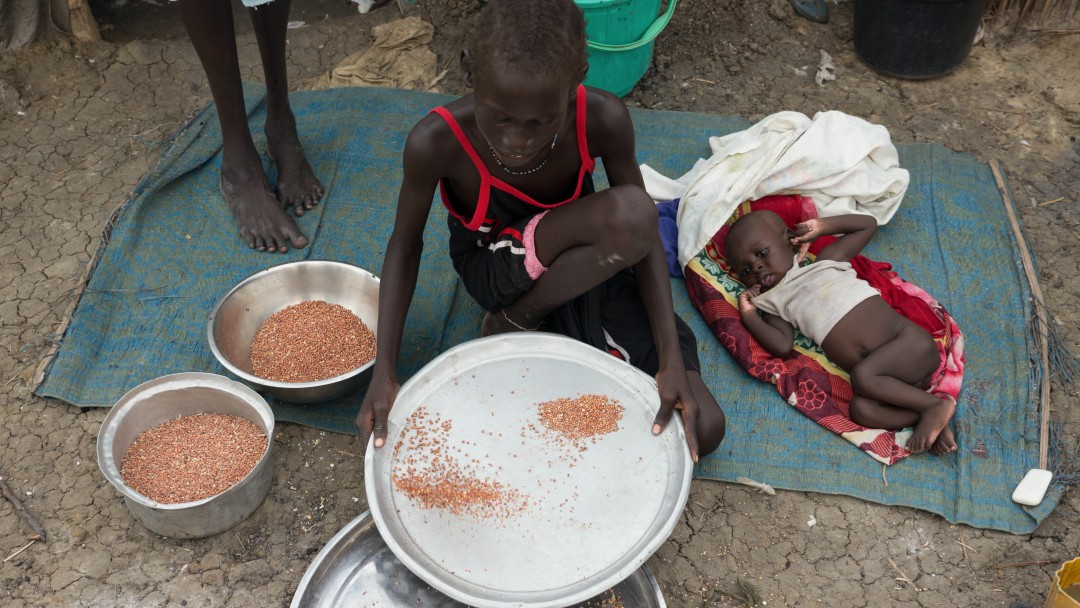 In einem Flüchtlingscamp im Südsudan trennt eine junge Mutter Spreu von Getreide, neben sich ihr Baby auf einer Matte.