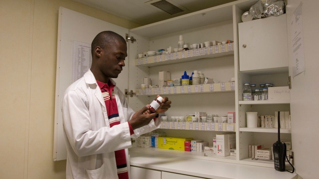 An African doctor looks at a medicine tin, other medicines are in a cupboard in the background. 