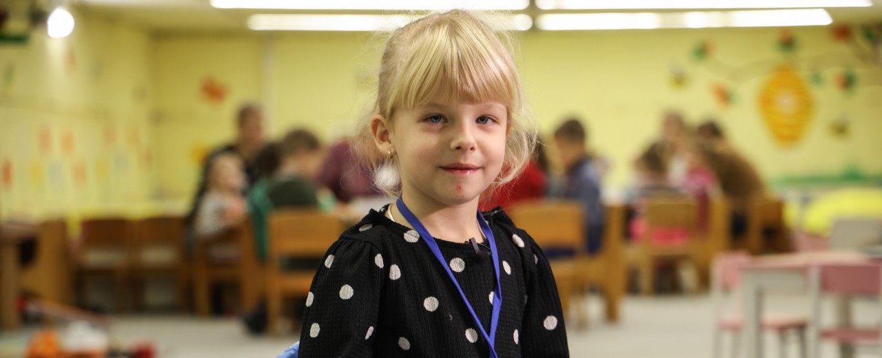 Blondes Mädchen mit Pferdeschwanz und Pony guckt in die Kamera. Im Hintergrund spielen Kinder an einem Tisch.