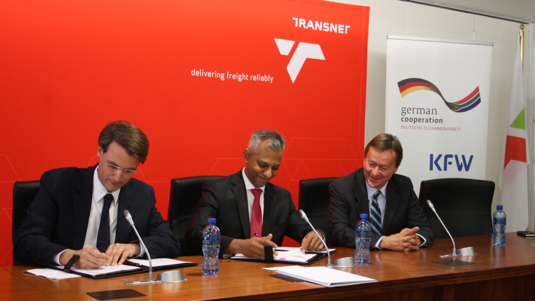(v.l.n.r.) Dr. Jan Martin Witte (Teamleiter Infrastruktur Südliches Afrika) Anoj Singh (CEO Transnet) und Dr. Horst Freitag (Deutscher Botschafter in Südafrika) bei der Vertragsunterzeichnung. Hinter ihnen sieht man die Logos der Organisationen
