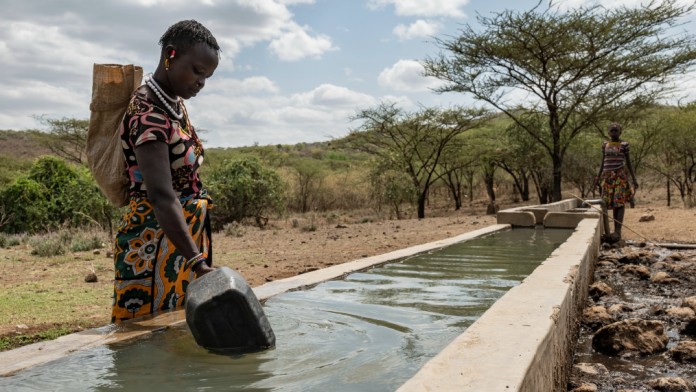 A traditionally dressed African woman fills a canister at a long water basin.