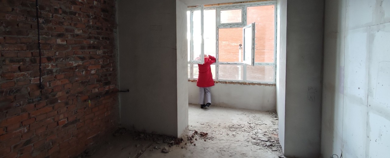 Eine Person steht in einem wiederaufgebautem Haus und schaut aus dem Fenster 