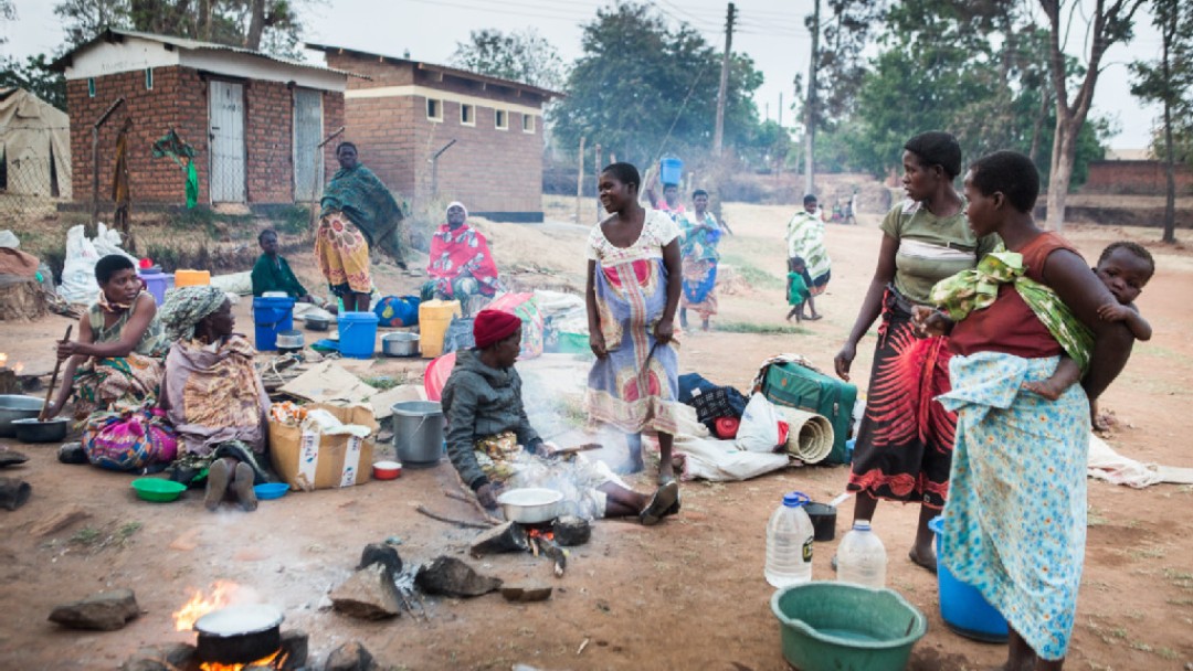 Pregnant women and women with their children are sitting in a courtyard outside of a clinic and prepare dinner on the ground.