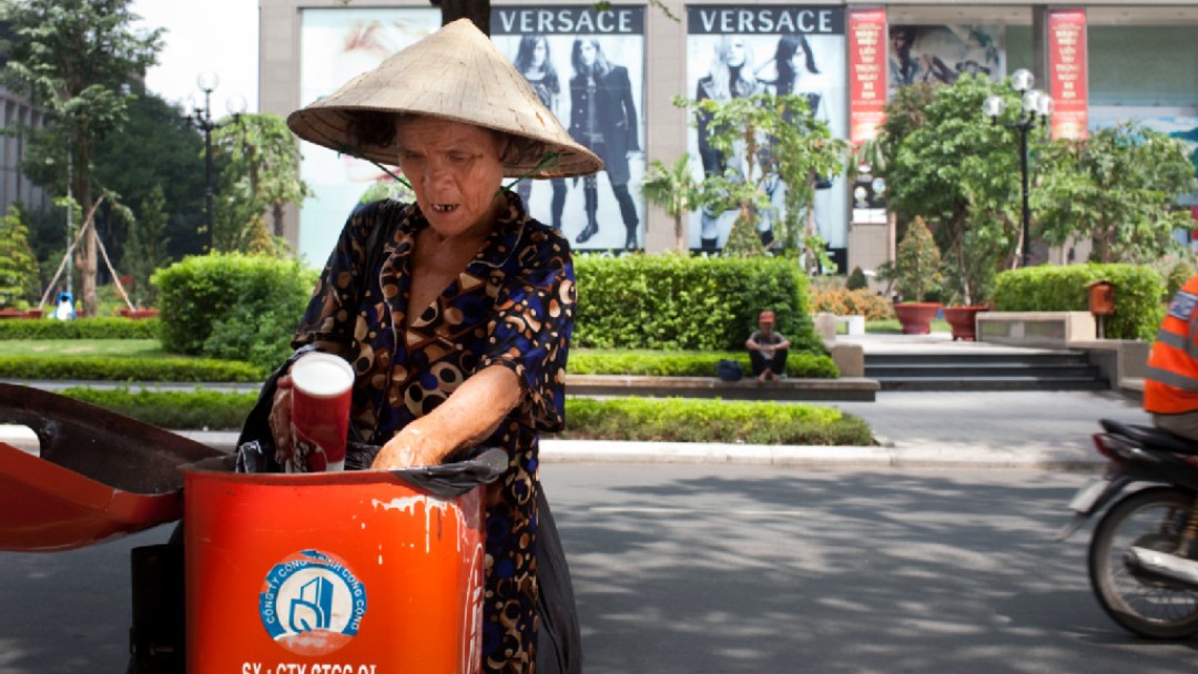 Frau mit Hut wühlt im Müll, im Hintergrund Versace