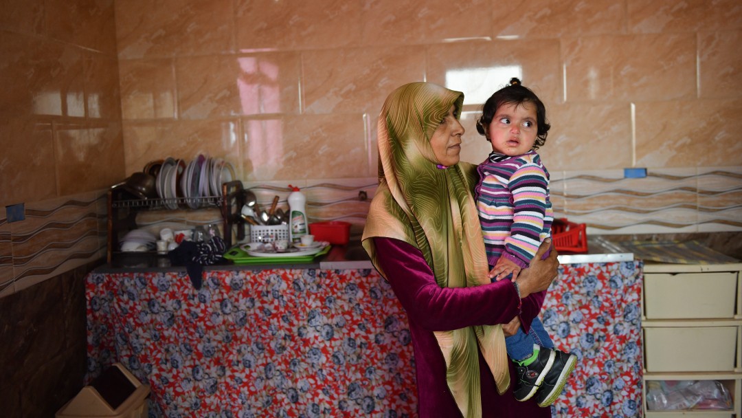 A woman carries a child, behind her a kitchenette