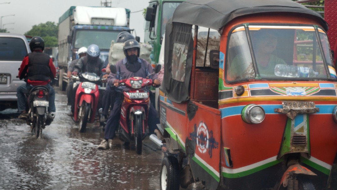 Ein Tuk-Tuk und mehrere Motorräder fahren auf einer nassen Straße in Asien.