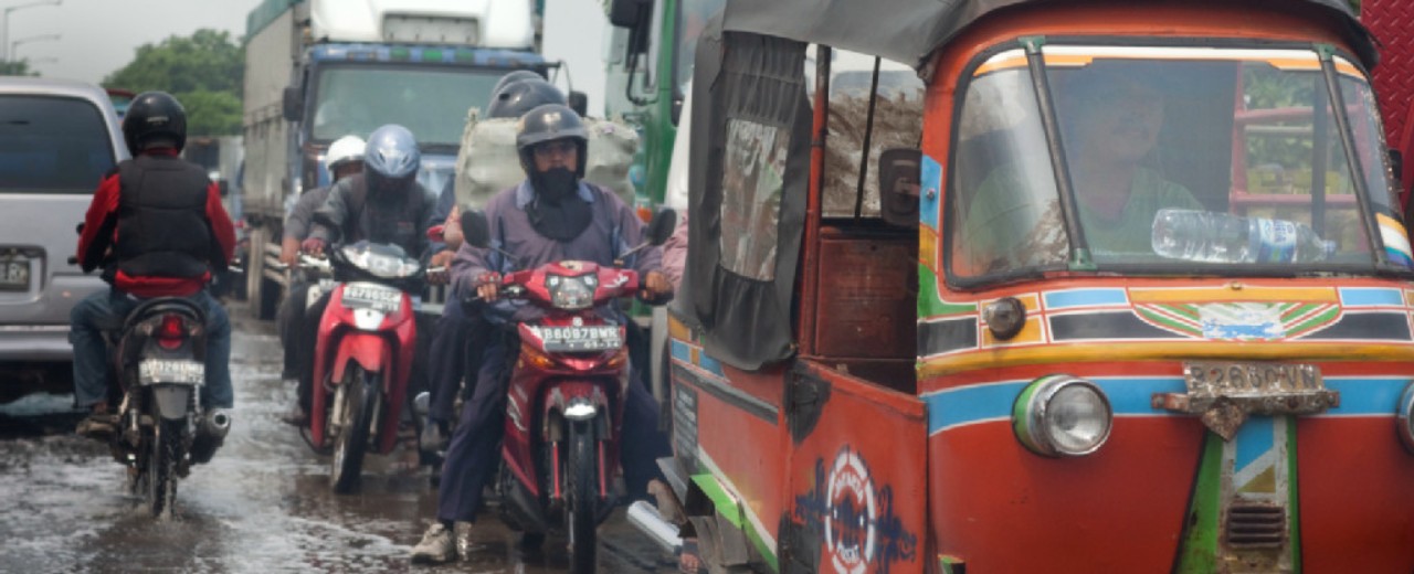 A Tuk-Tuk and several motorbikes are driving on a wet road in Asia.