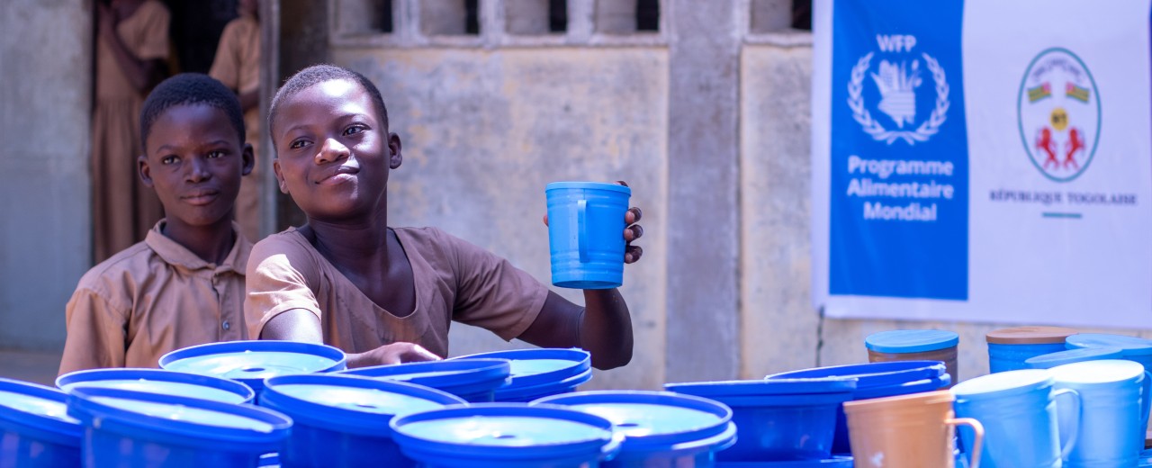 Two schoolchildren holding a jug, standing in front of several buckets