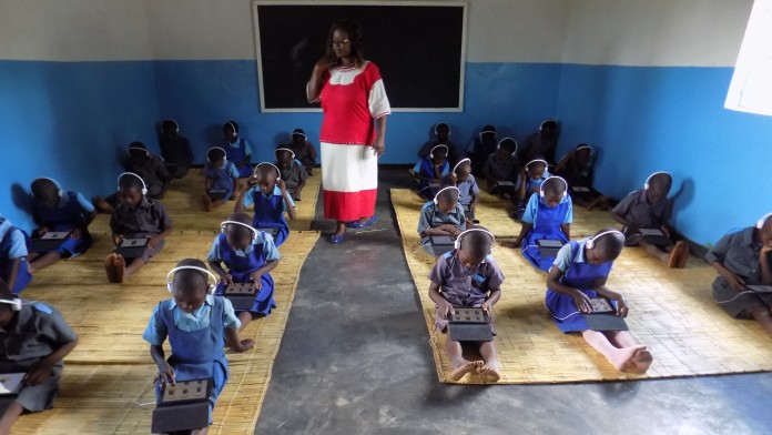 Malawi: Students sit on the floor in the classroom with trays on their laps, on which they solve school tasks, observed by their teacher.