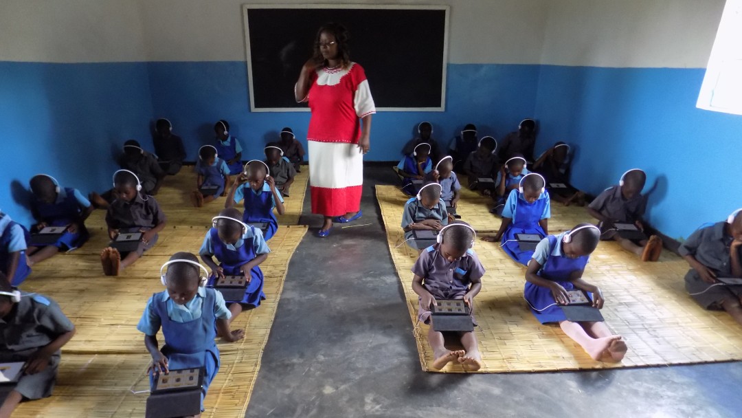 Malawi: Schüler sitzen im Klassenraum auf dem Boden mit Tablets auf ihren Schößen, auf denen sie, von ihrer Lehrerin beobachtet, Schulaufgaben lösen.