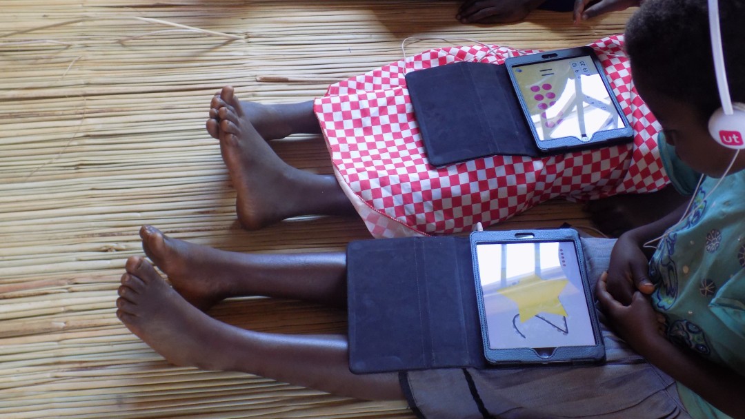 Malawi: two pupils sit on the floor in the classroom with a tablet on their laps on which they solve arithmetical problems.