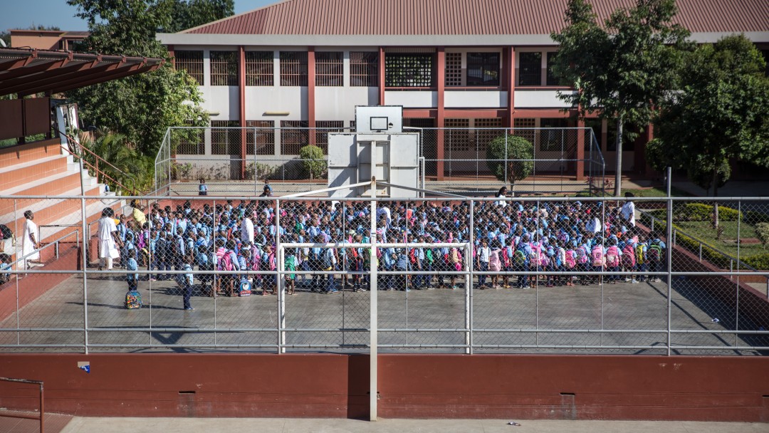Primary school in Maputo: three teachers gather the pupils on the school's sports field with the school building in the background.