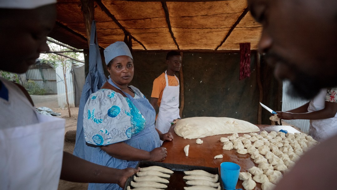 A woman stands at a table and forms dough into bread. Other men and women join in.