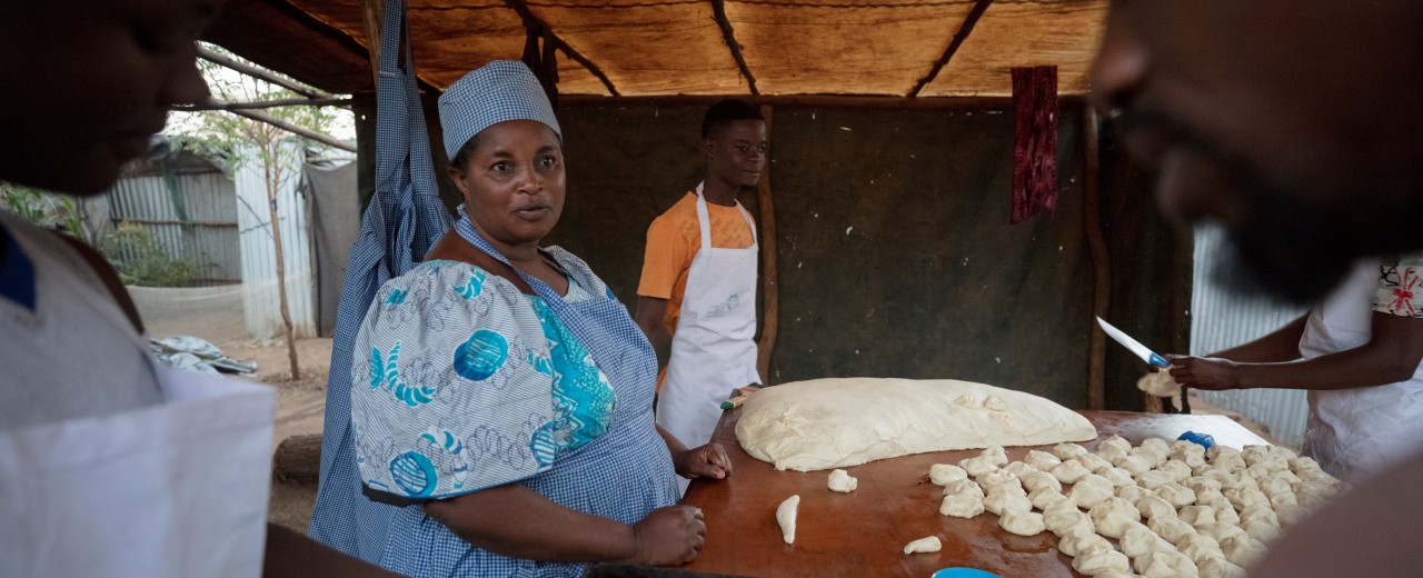 A woman stands at a table and forms dough into bread. Other men and women join in.