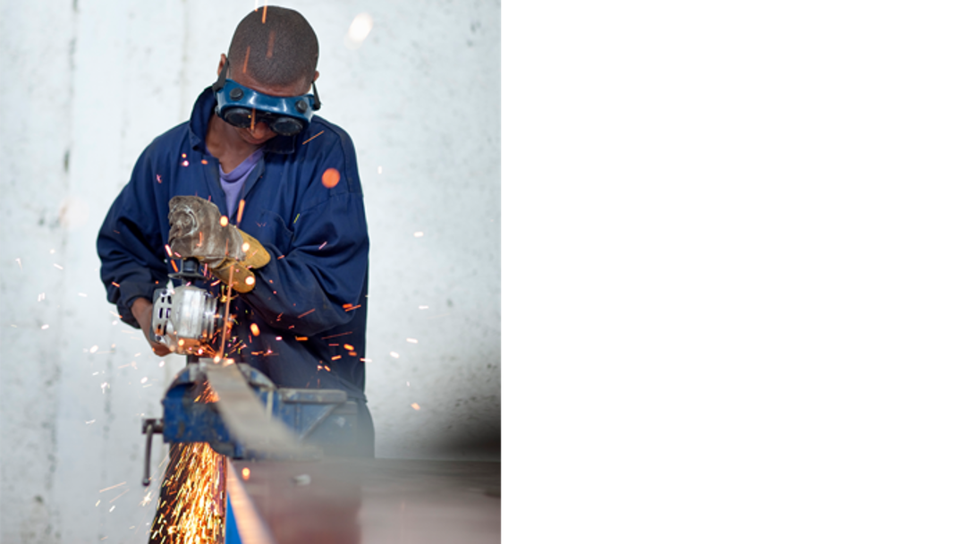 A young African worker wearing protective goggles grinds a plate, causing sparks to fly