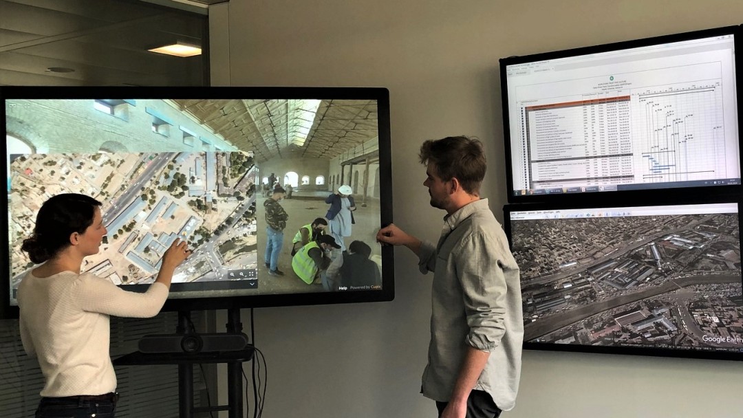 Room with several screens, on one of them two colleagues show pictures from a construction site, on the other one you see the floor plan, some statistics and other colleagues watching. 