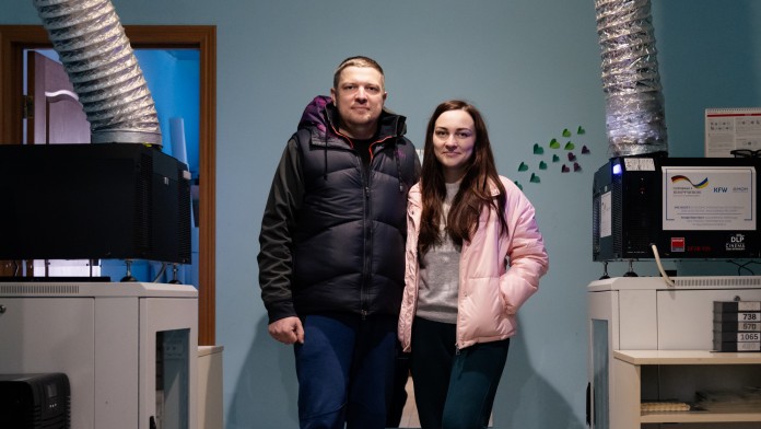 Ein junger Mann und eine junge Frau stehen in einem Raum mit technischen Geräten. Zwei Belüftungsröhre führen zur Decke.