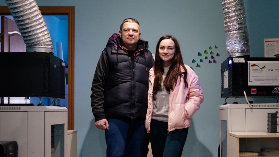 Ein junger Mann und eine junge Frau stehen in einem Raum mit technischen Geräten. Zwei Belüftungsröhre führen zur Decke.