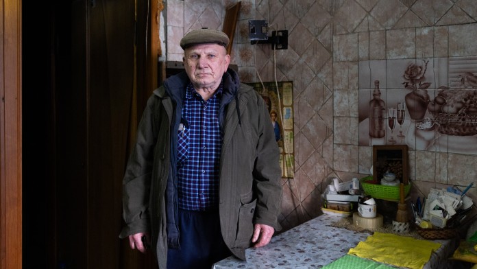 Ein älterer Mann mit Kappe und dicker Jacke steht an einem Tisch in einer deckenhoch gekachelten Küche. Oberhalb des Tisches befindet sich ein gekacheltes Bild.
