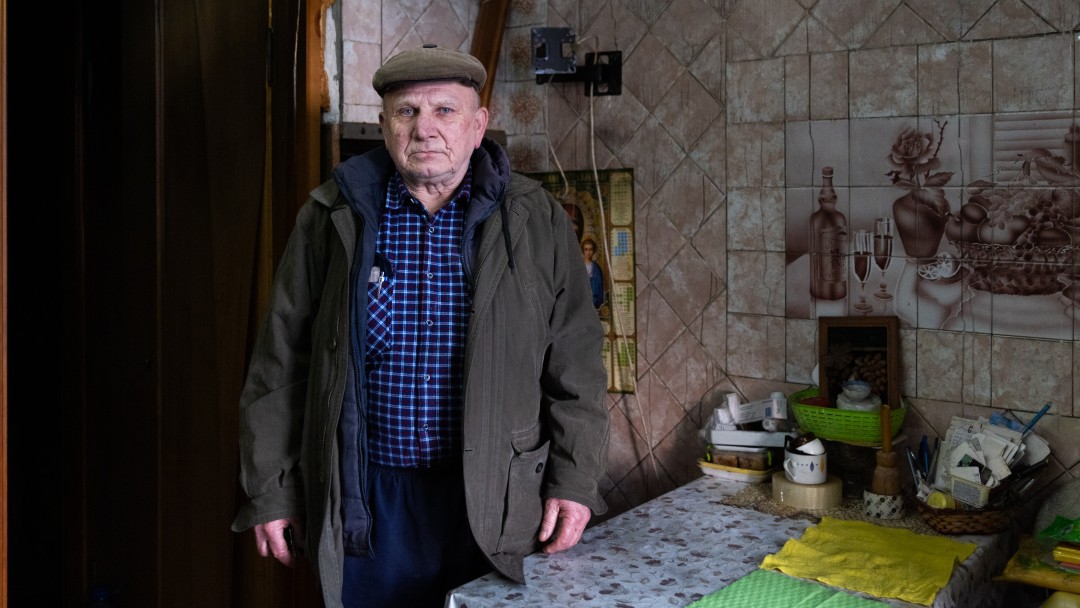 Ein älterer Mann mit Kappe und dicker Jacke steht an einem Tisch in einer deckenhoch gekachelten Küche. Oberhalb des Tisches befindet sich ein gekacheltes Bild.