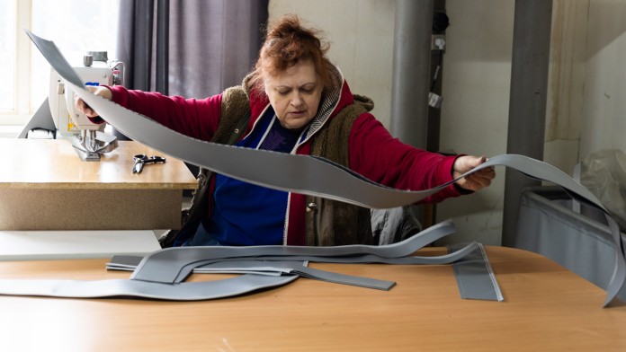 Eine Frau breitet auf einem Tisch zugeschnittene Lederteile aus. Im Hintergrund ist eine Nähmaschine zu sehen.