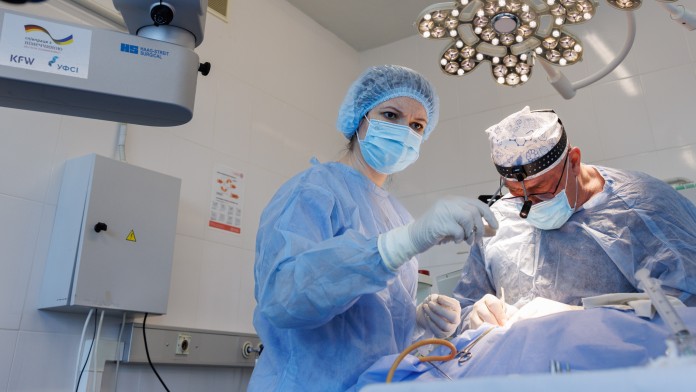 Zwei Ärzte in blauer OP-Kleindung beugen sich über den OP-Tisch. Im Vordergrund ist ein technisches Gerät zu sehen.