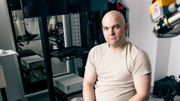 Ein Mann mit Glatze sitzt auf einem Trainingsgerät und hält seinen versehrten rechten Fuß hoch.