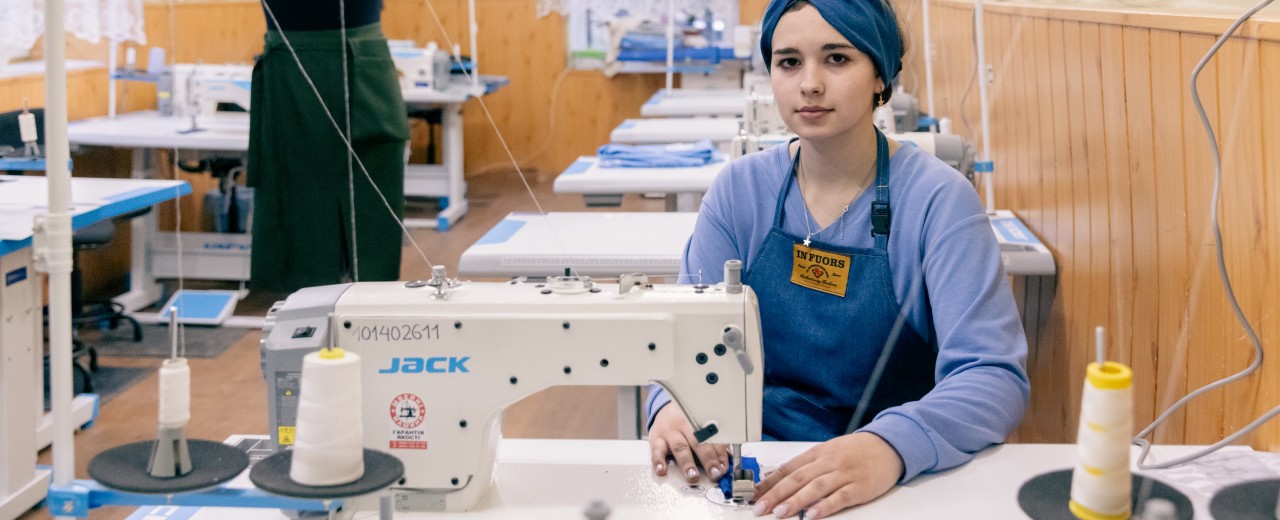 A young woman is sitting at a sewing machine, in the background there are other workstations, a dressmaker's dummy and a blackboard and screen on the wall.