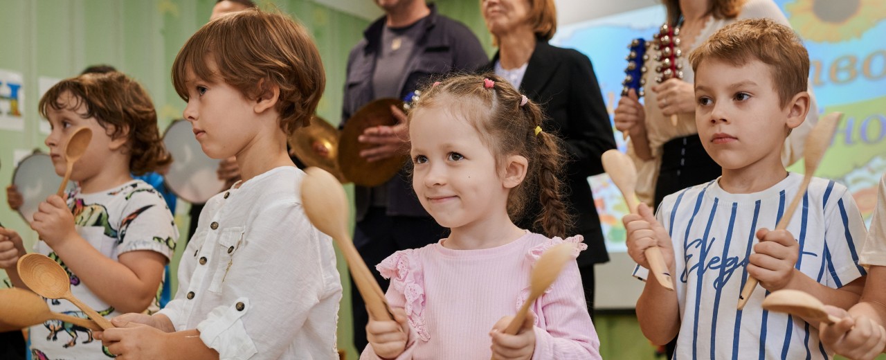 Children stand in a row with cooking spoons as musical instruments