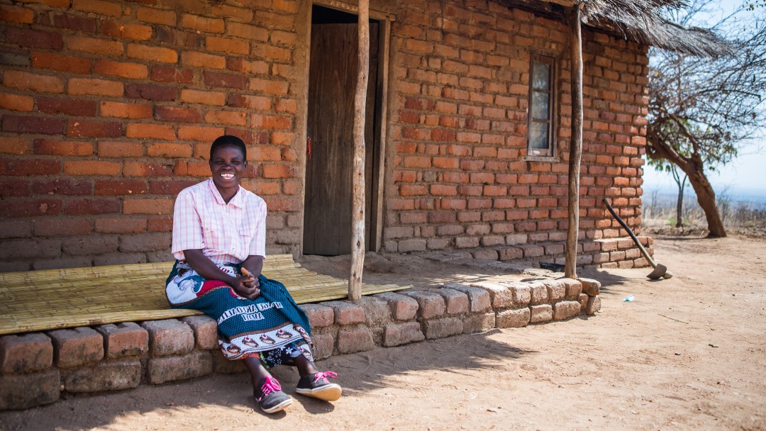 Woman sitting in front of a stone hut. She is participating in the "Social Cash Transfer" programme